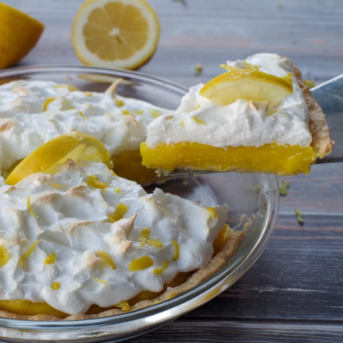 a piece of lemon pie being lifted out of a whole pie, with a lemon cut in half in background