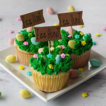 3 Mini egg cupcakes on a square white plate with more mini eggs and candy flowers scattered around