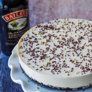 Whole No bake Baileys cheesecake on a white cake platter with a bottle of baileys in the background