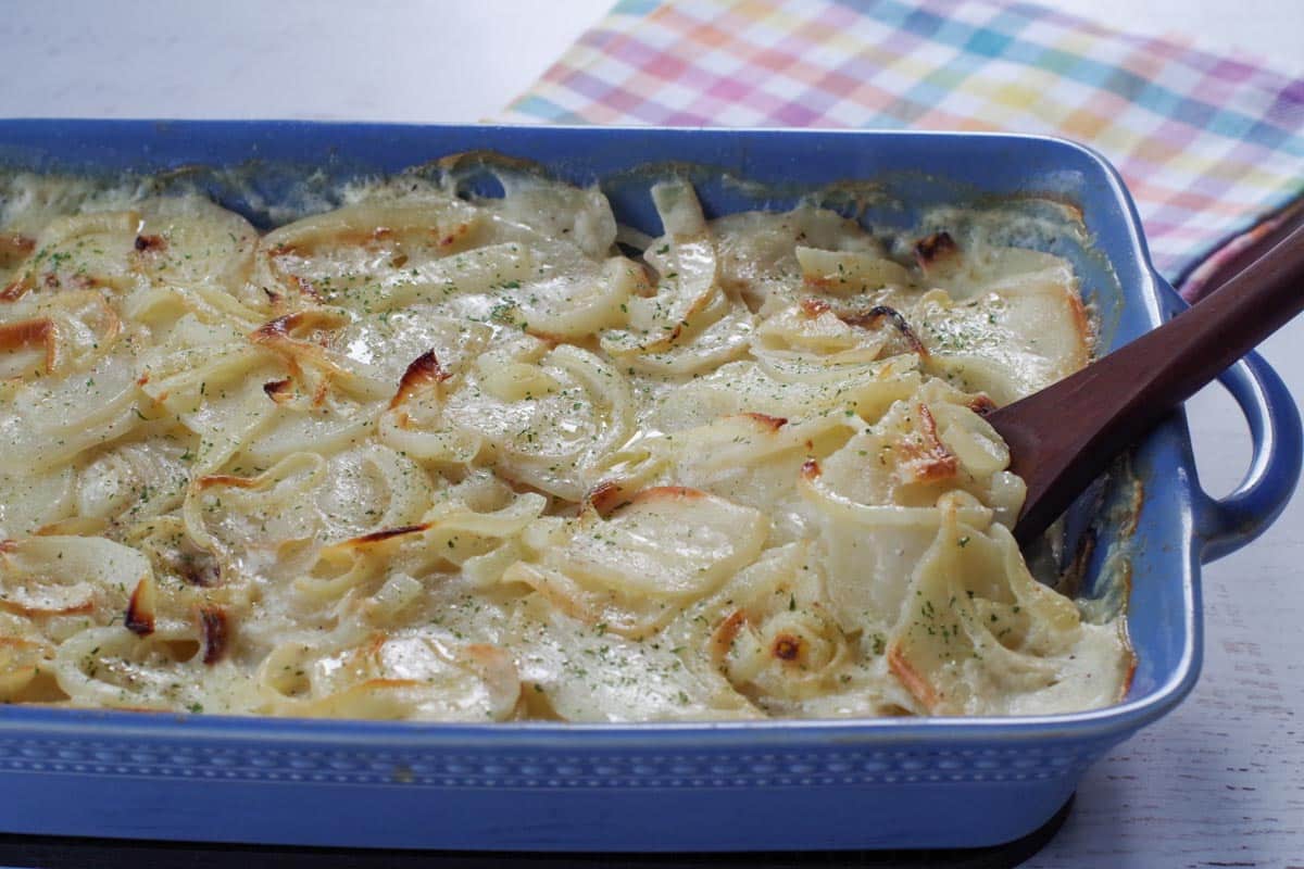 cooked scalloped potatoes without cheese in a blue casserole dish with a wooden spoon and a checkered tea towel in the background