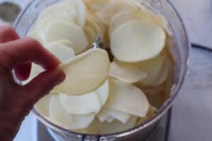 hand holding up a slice of potato over a food processor with more sliced potatoes in bowl