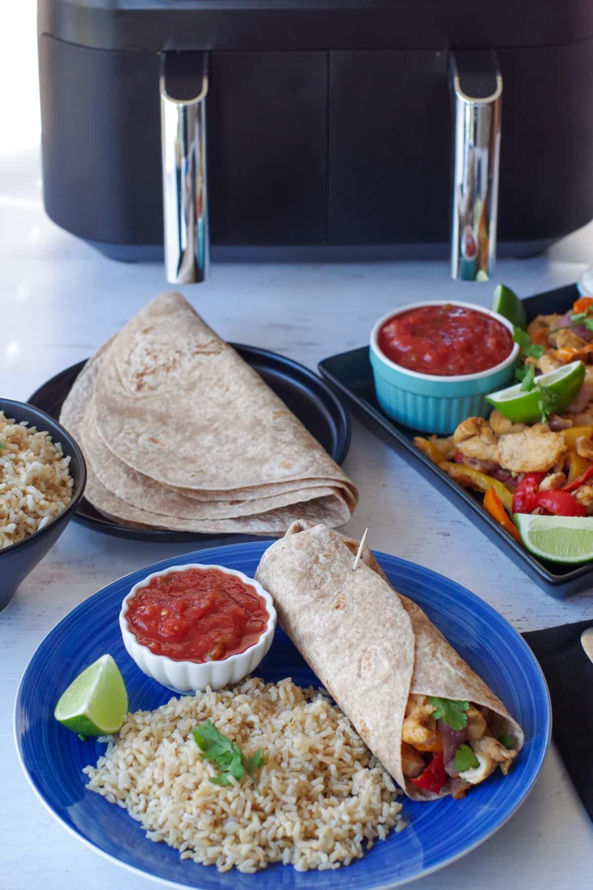a blue plate with a fajita wrap and rice, then a tray of fajita mix, tortillas, rice and a Ninja foodi air fryer in the background