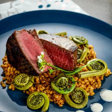 beef tenderloin with wheat berries and fiddleheads on a blue plate