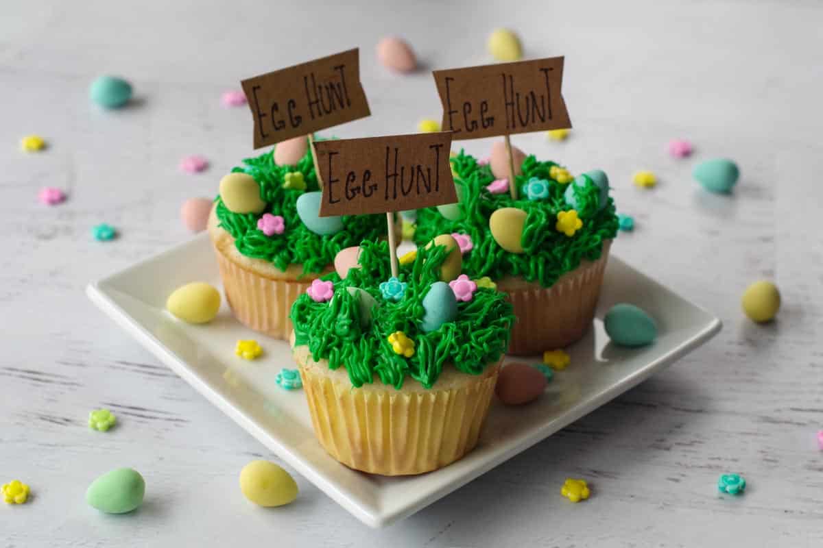 3 Mini egg cupcakes on a square white plate with more mini eggs and candy flowers scattered around