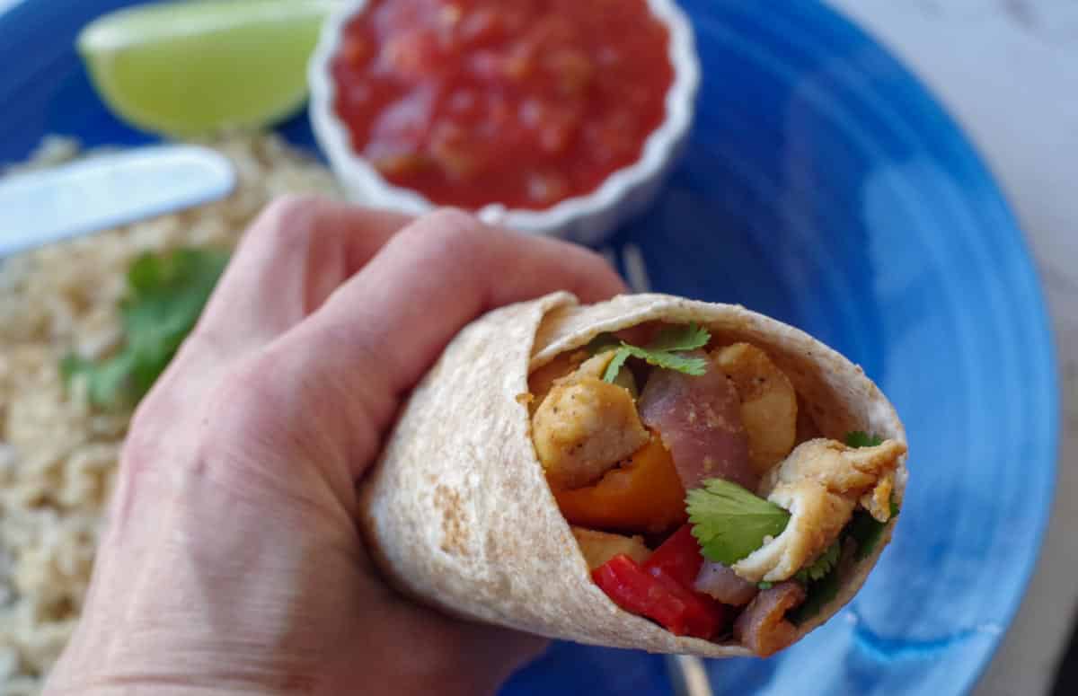hand holding up a chicken fajita wrap over a blue plate with salsa
