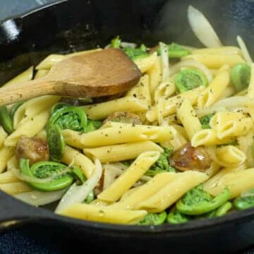 fiddlehead sausage penne in a cast iron frying pan, with a wooden spoon stirring it