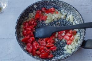 tomatoes added to frying pan with onion and garlic, with a black spatula