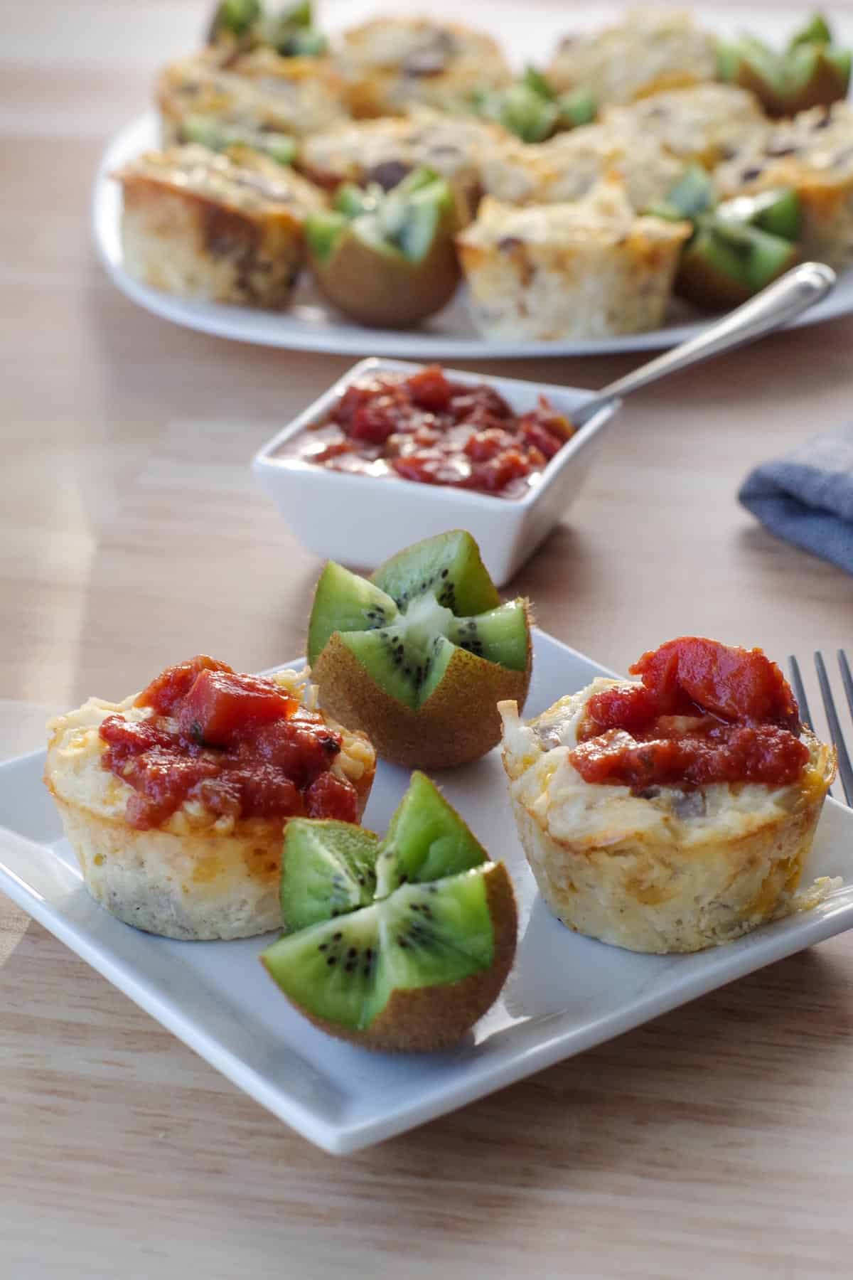2 brunch bites with salsa on top, on a small white plate with kiwi flowers and salad in background and fork on righthand side