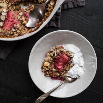 baked rhubarb oatmeal in a white dish with a spoon (on black surface) with container of rhubarb oatmeal in the background