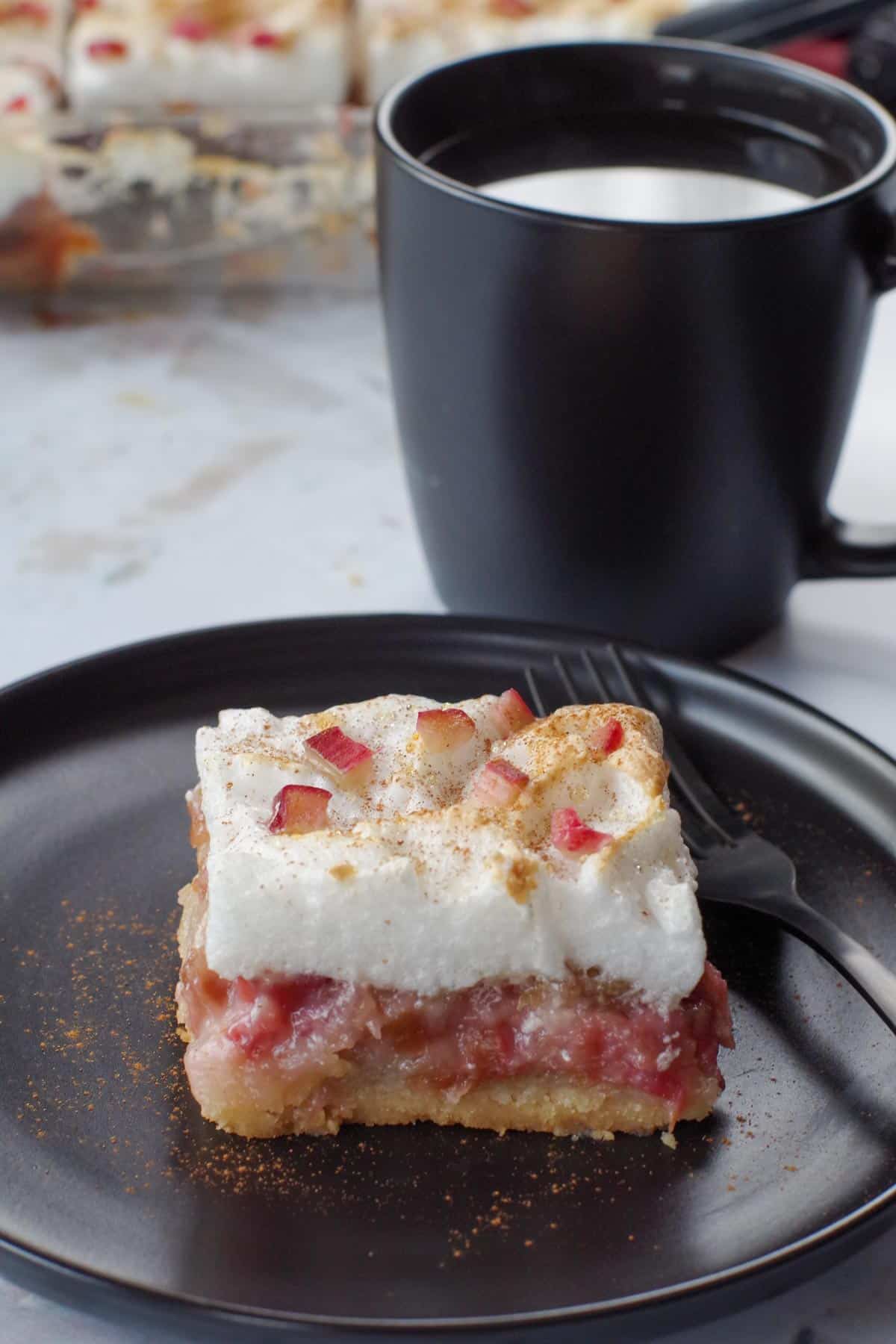 a piece of rhubarb meringue torte on a black plate with a fork and a black cup of coffee in background