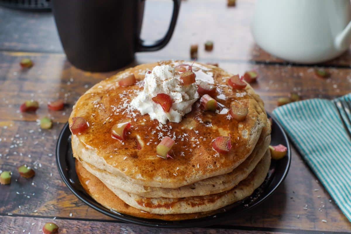 4 Rhubarb pancakes stacked with garnish of whipped cream, coconut, cinnamon and syrup on black plate, with black coffee cup and white syrup container in background