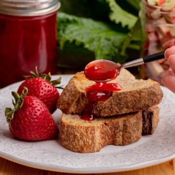 rhubarb jam on a spoon being spread over toast, on a white plate, with strawberries and container of jam in the background