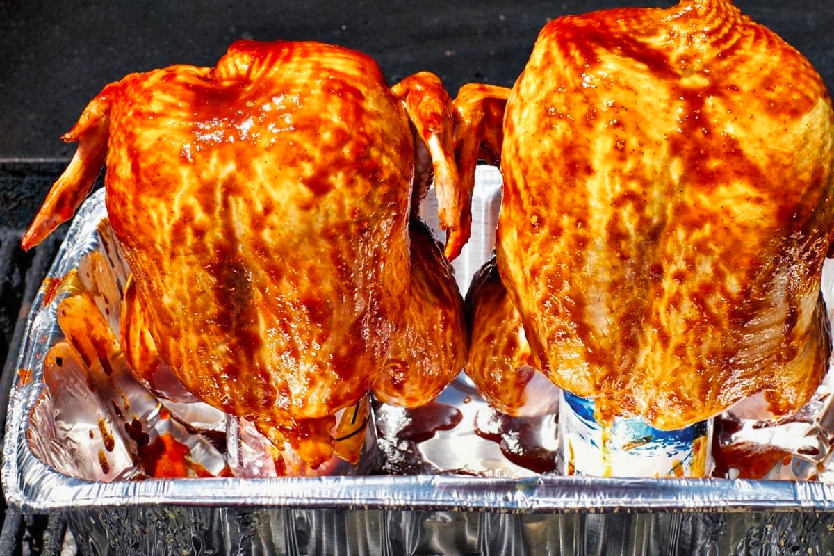2 beer butt chickens with bbq sauce on cans of beer in an aluminum container