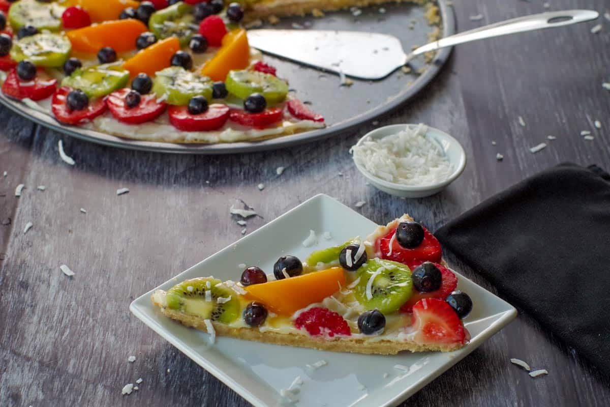 piece of fruit pizza on a white plate with whole fruit pizza in the background