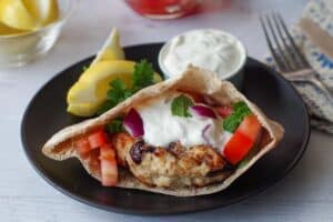 Greek chicken burger in a pita on a black plate with lemon wedge and small dish of tzatziki