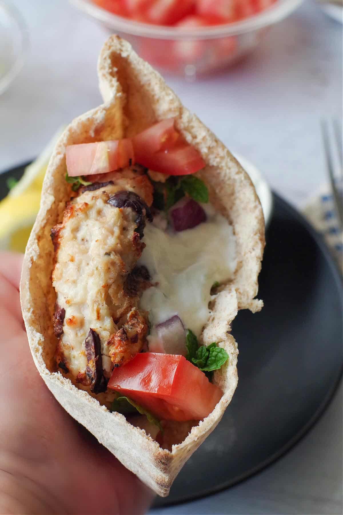 Greek chicken burger in a pita, held up by hand, over a black plate