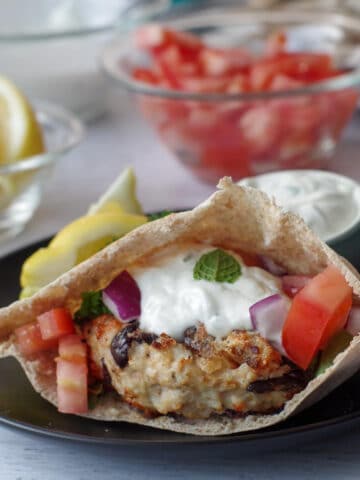 Greek chicken burger in a pita on a black plate with lemon wedge and small dish of tzatziki