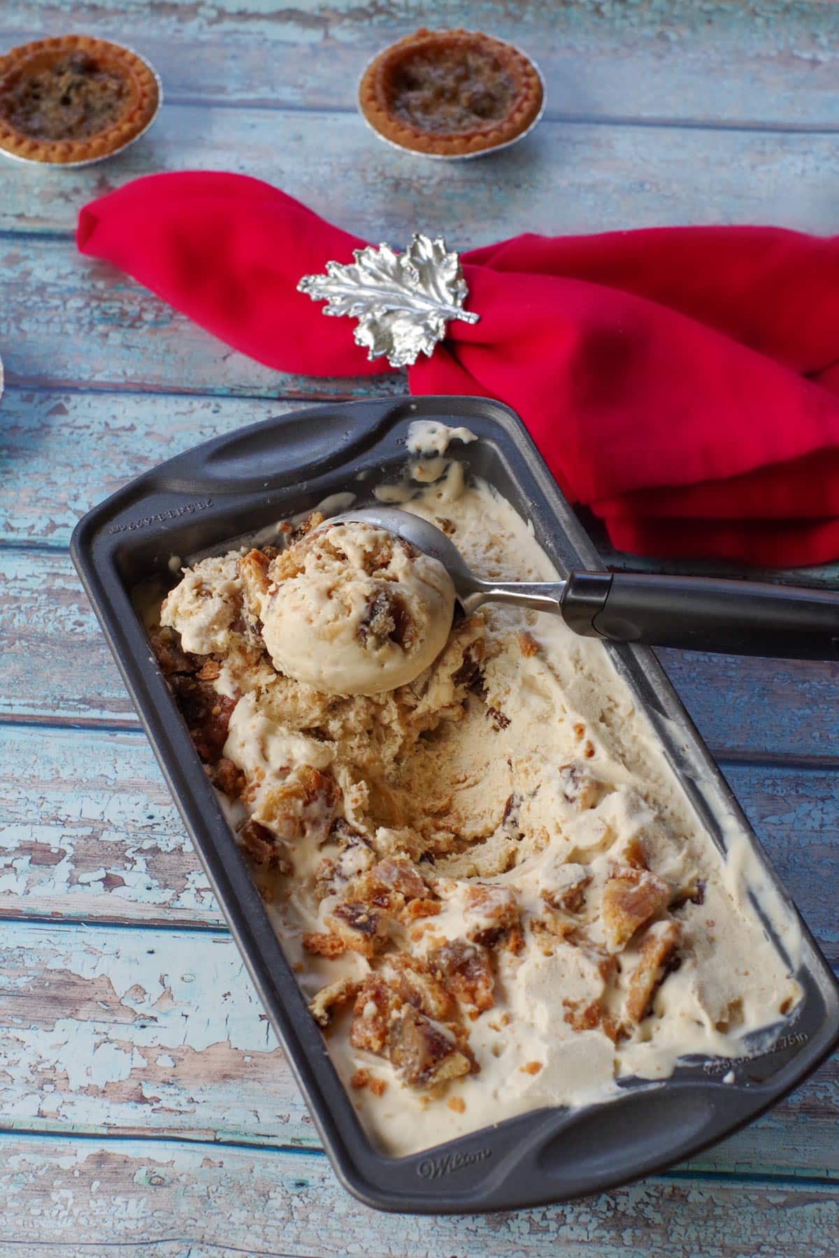 butter tart ice cream in a loaf pan being scooped out with ice cream scoop a red napkin with silver maple leaf napkin ring and butter tarts in the background