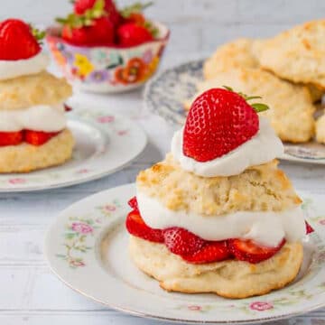 2 strawberry shortcakes on white plates, with a bowl of strawberries in background