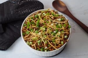 chow mein casserole garnished with green onion and dry noodles, with wooden spoon on one side, and black oven mitts on other side