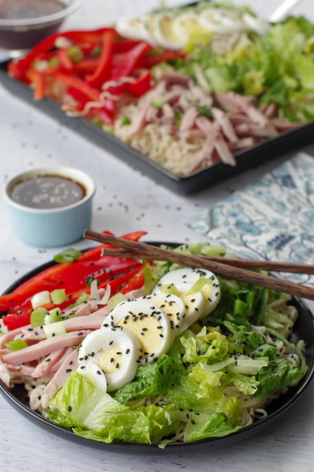Japanese cold noodle salad on a black plate, with chopsticks and a dish of dressing in the background, as well as a black serving tray with more salad