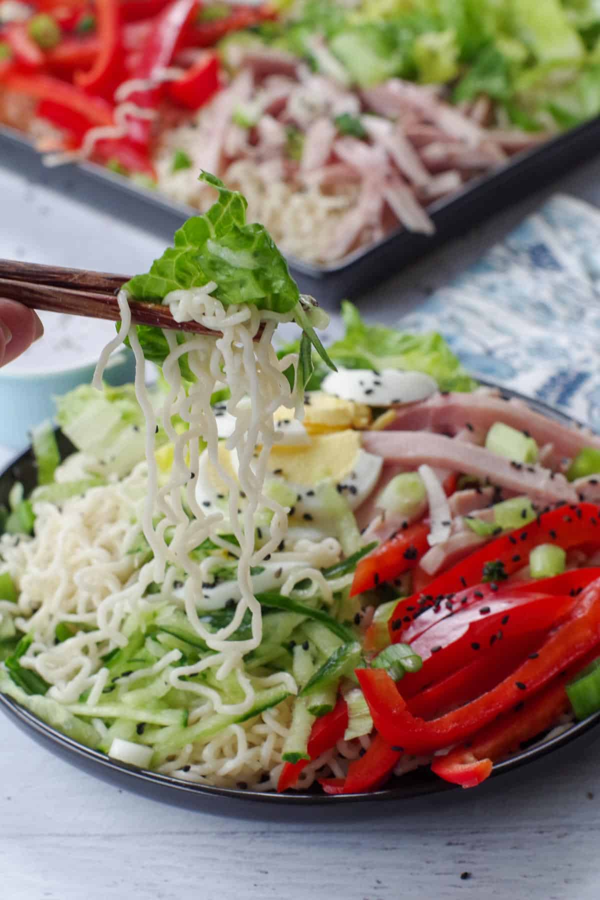 cold noodle salad being lifted up with chopsticks, over a black round plate, with a black serving tray of more salad in backgrond