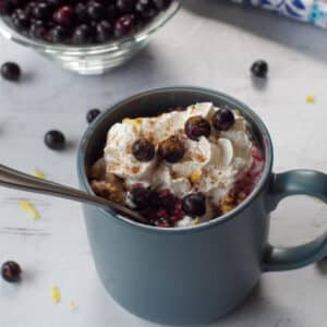 Saskatoon berry crisp recipe in a mug with a spoon in it, berries stewn around and a bowl of Saskatoon berries in the background