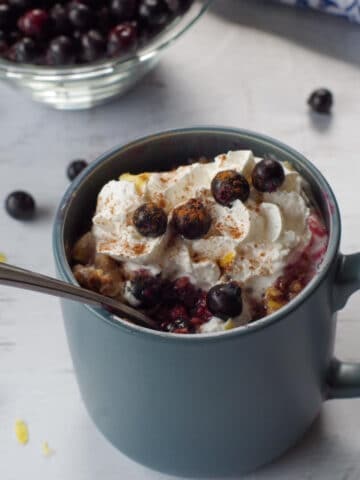 Saskatoon berry crisp recipe in a mug with a spoon in it, berries stewn around and a bowl of Saskatoon berries in the background