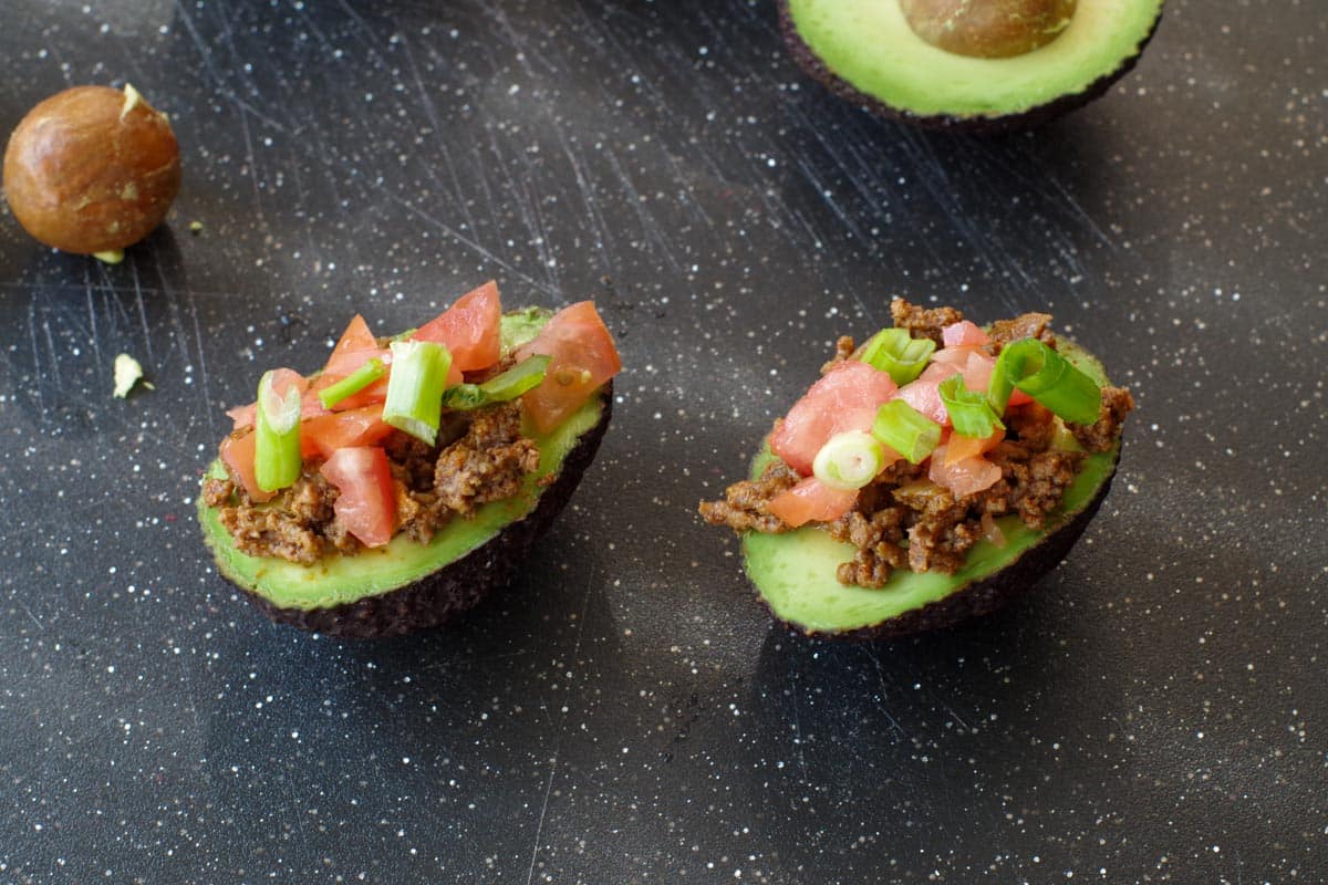 2 avocado tacos on black cutting board with ground beef, tomatoes and onion added