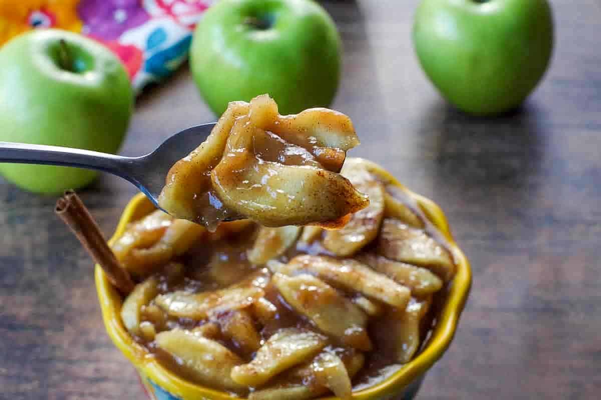 apple pie filling being held up on spoon over bowl of more pie filling, with apples in background