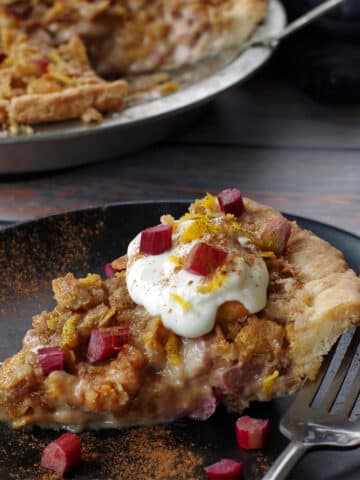 a slice of sour cream rhubarb pie on a black plate with a fork on the plate and whole remaining pie in the background
