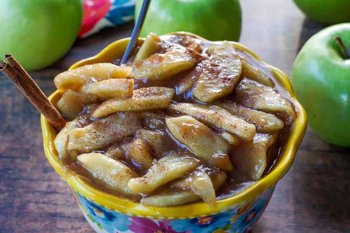 stovetop apple pie filling in a flowered blue bowl with yellow rim, with spoon and cinnamon stick in it