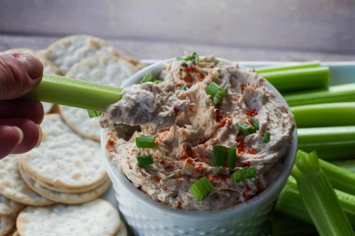 tuna dip in white container on a tray with crackers and celery with piece of celery being dipped into the tuna dip