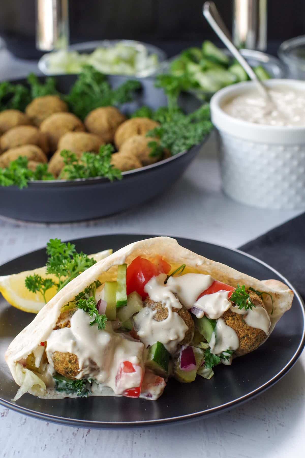 air fryer falafel in pita on black plate, with bowl of falafel balls and pita in the background