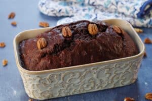 Entire loaf of chocolate zucchini loaf with blue patterned oven mitts in the background and pecans strewn about