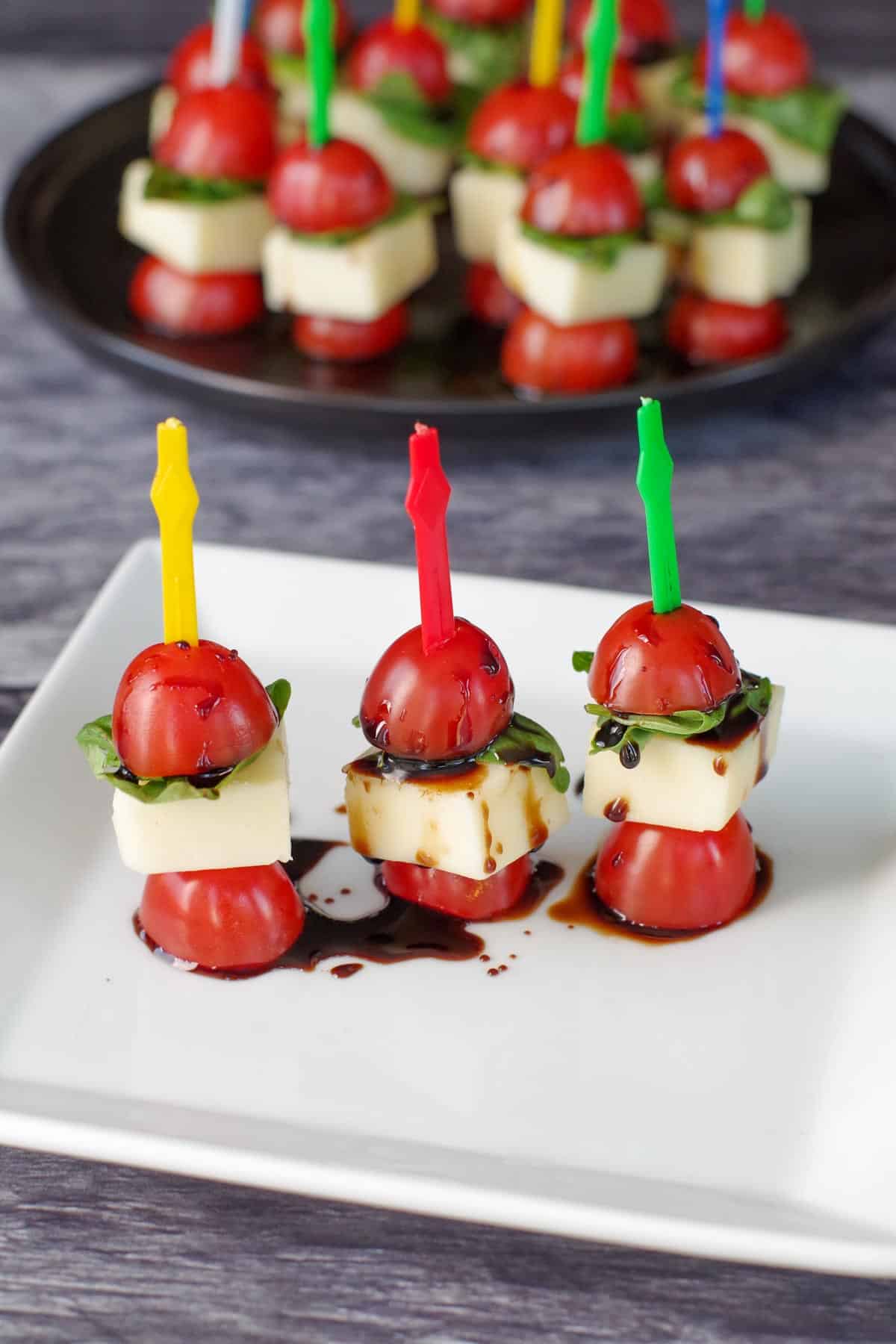 3 Caprese salad sticks on a white plate with a platter in the background