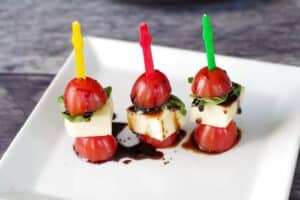 3 Caprese salad sticks on a white plate, drizzled with balsamic glaze