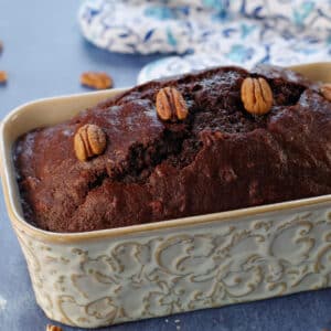 Entire loaf of chocolate zucchini loaf with blue patterned oven mitts in the background and pecans strewn about