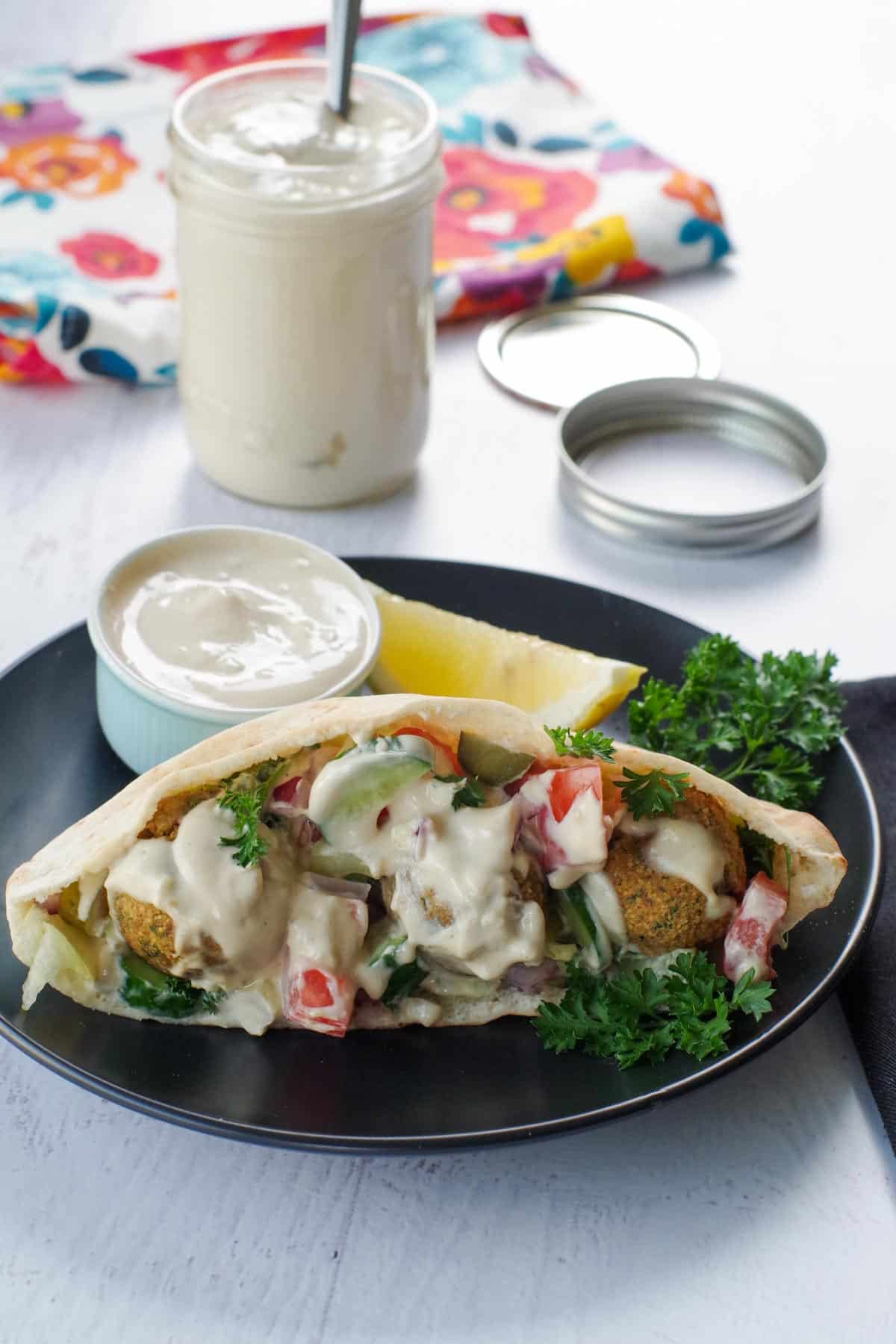 A falafel on a black plate, with a dish of tahini sauce and a jar of tahini sauce in the background