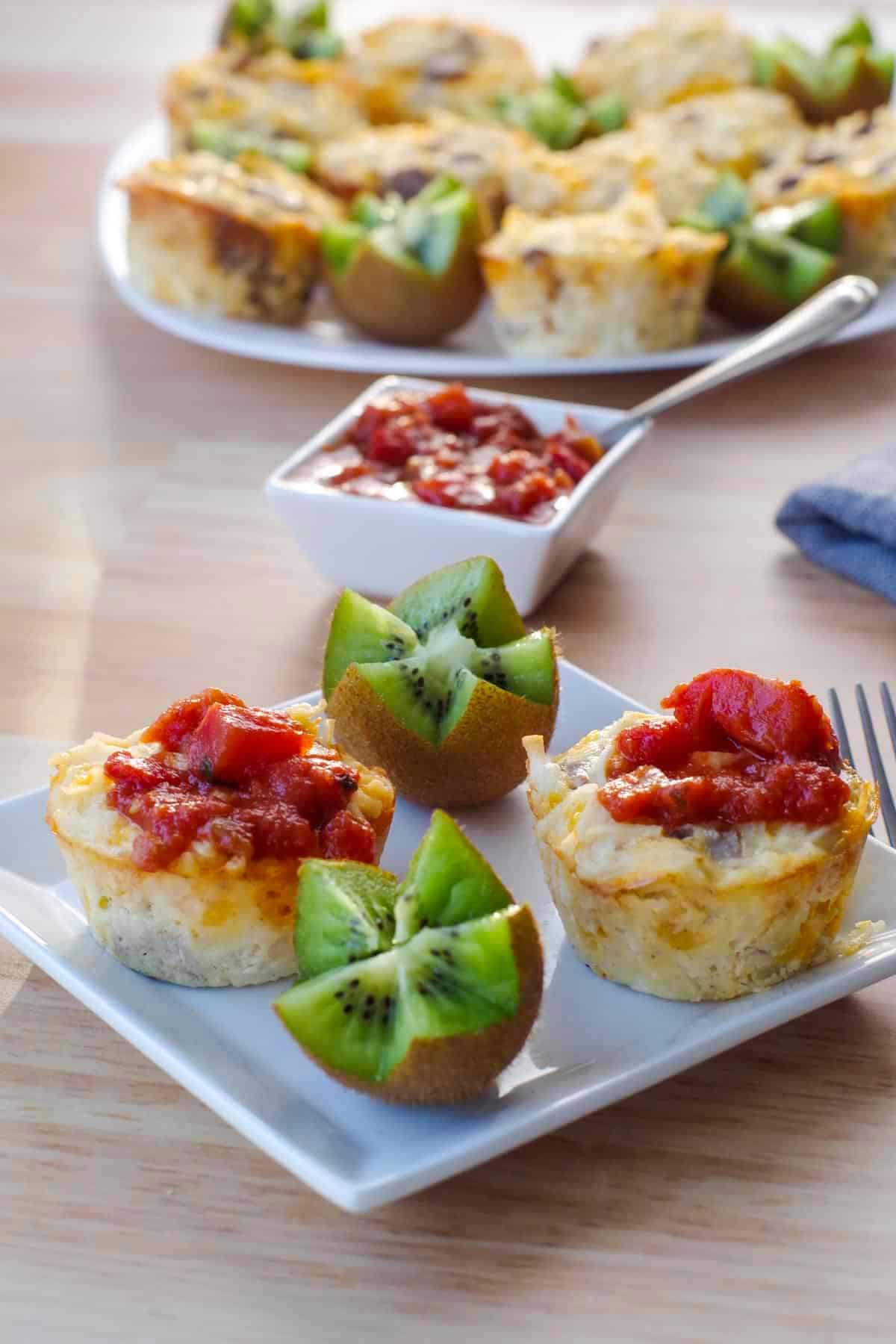 2 brunch bites with salsa on top, on a small white plate with kiwi flowers and salad in background and fork on righthand side and plate of more brunch bites in the background