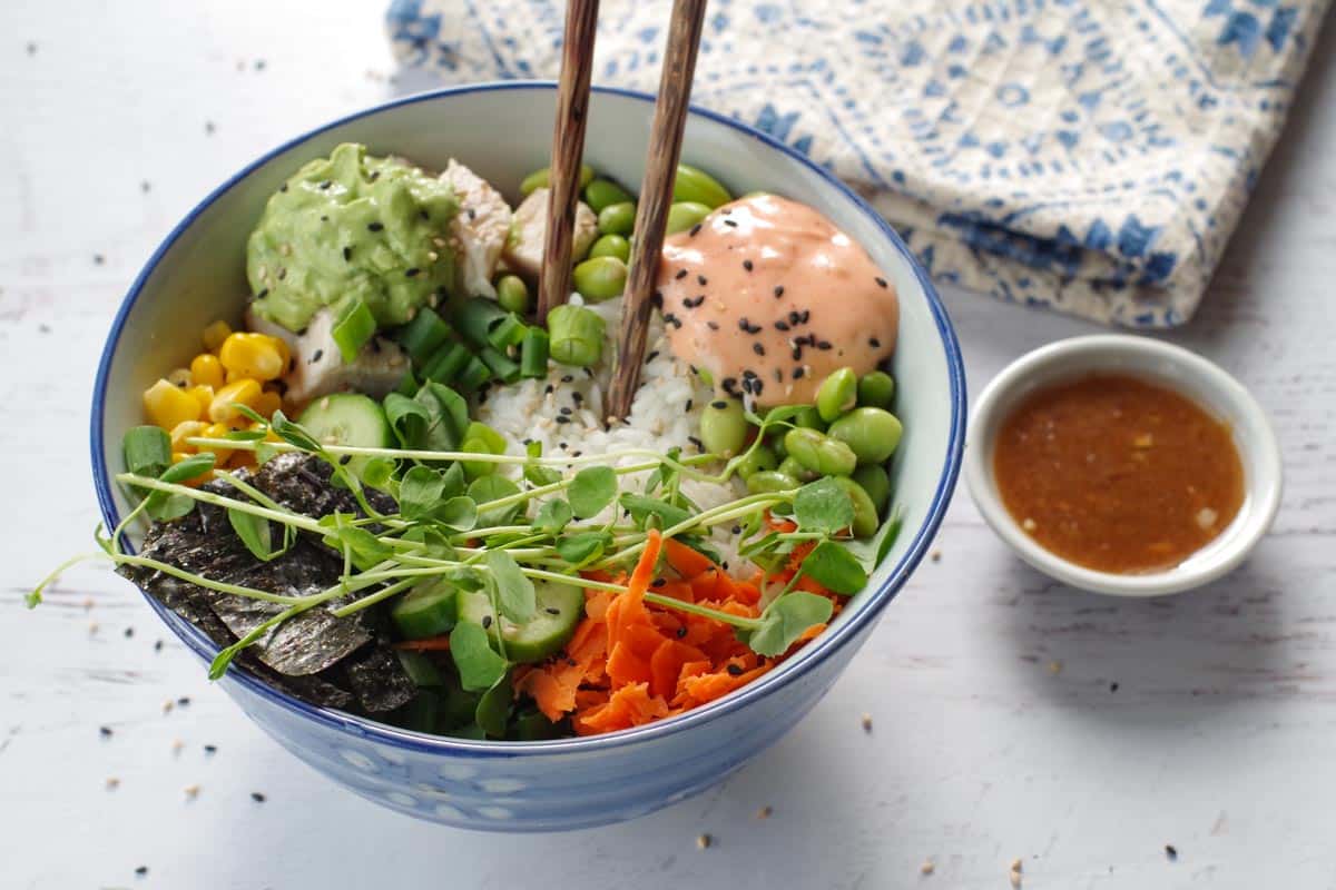 chicken poke bowl with chopsticks sticking out of bowl, small white dish of miso salad dressing on the side and white and blue patterned tea towel in the background
