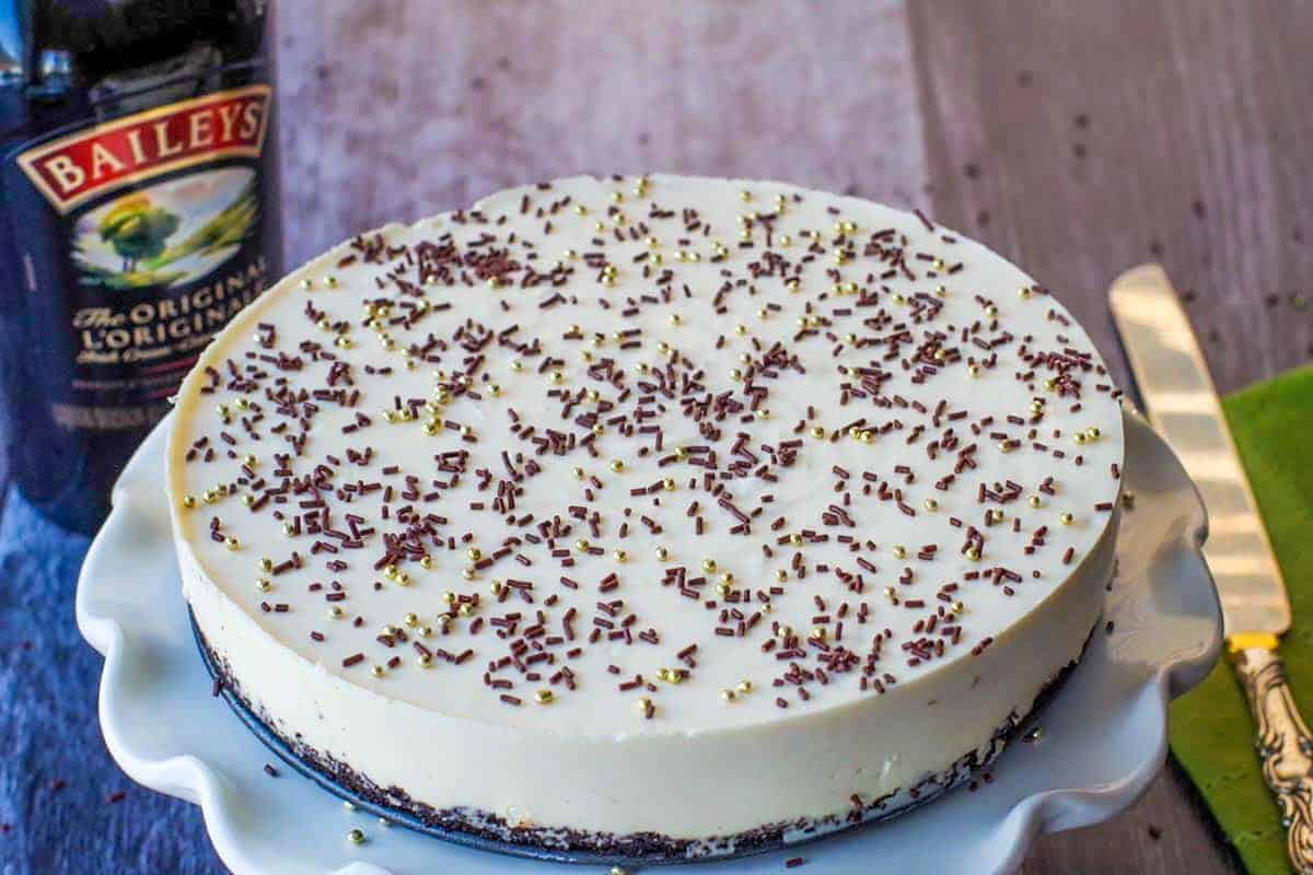 Whole No bake Baileys cheesecake on a white cake platter with a bottle of baileys in the background on the left and a knife on the right