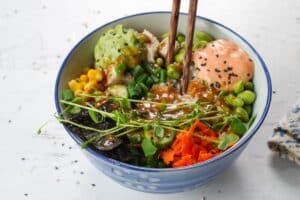 chicken poke bowl with micro greens, sauces, sesame seeds and dressing added, in a bowl with chopsticks.