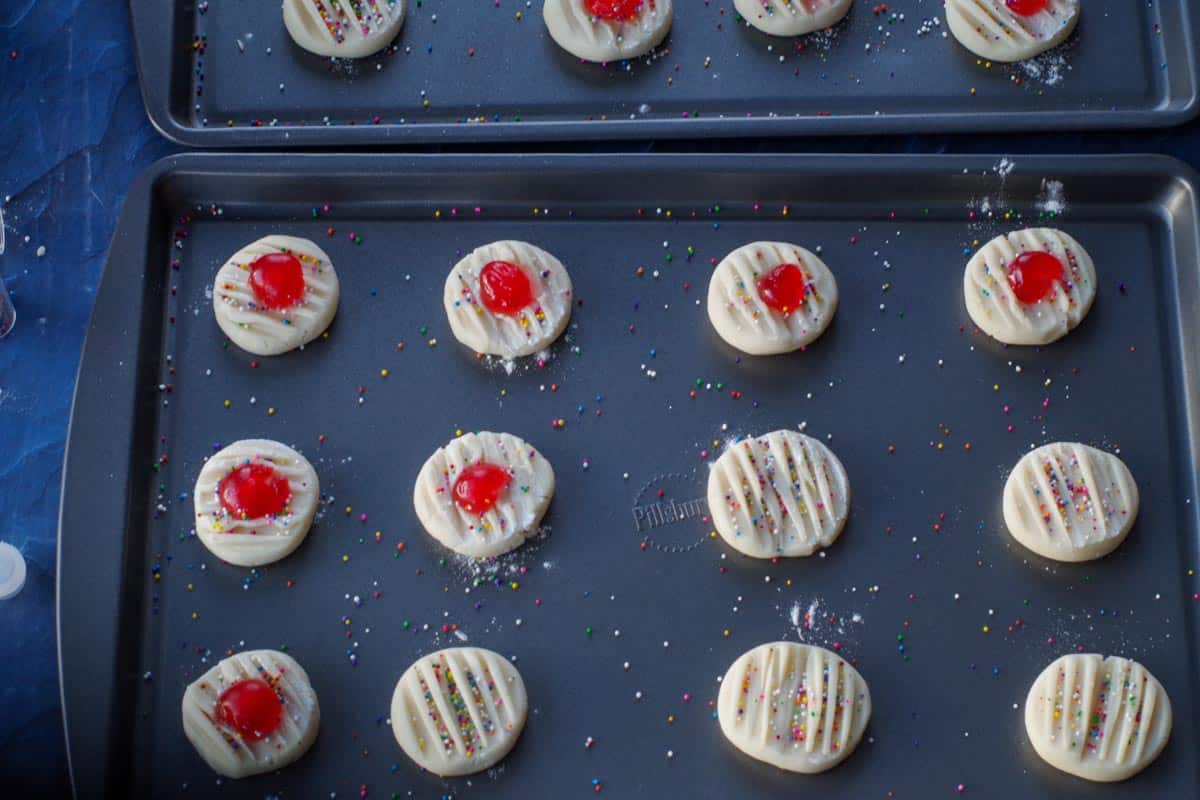 sprinkles and glazed cherry halves added to uncooked cookies on baking sheet