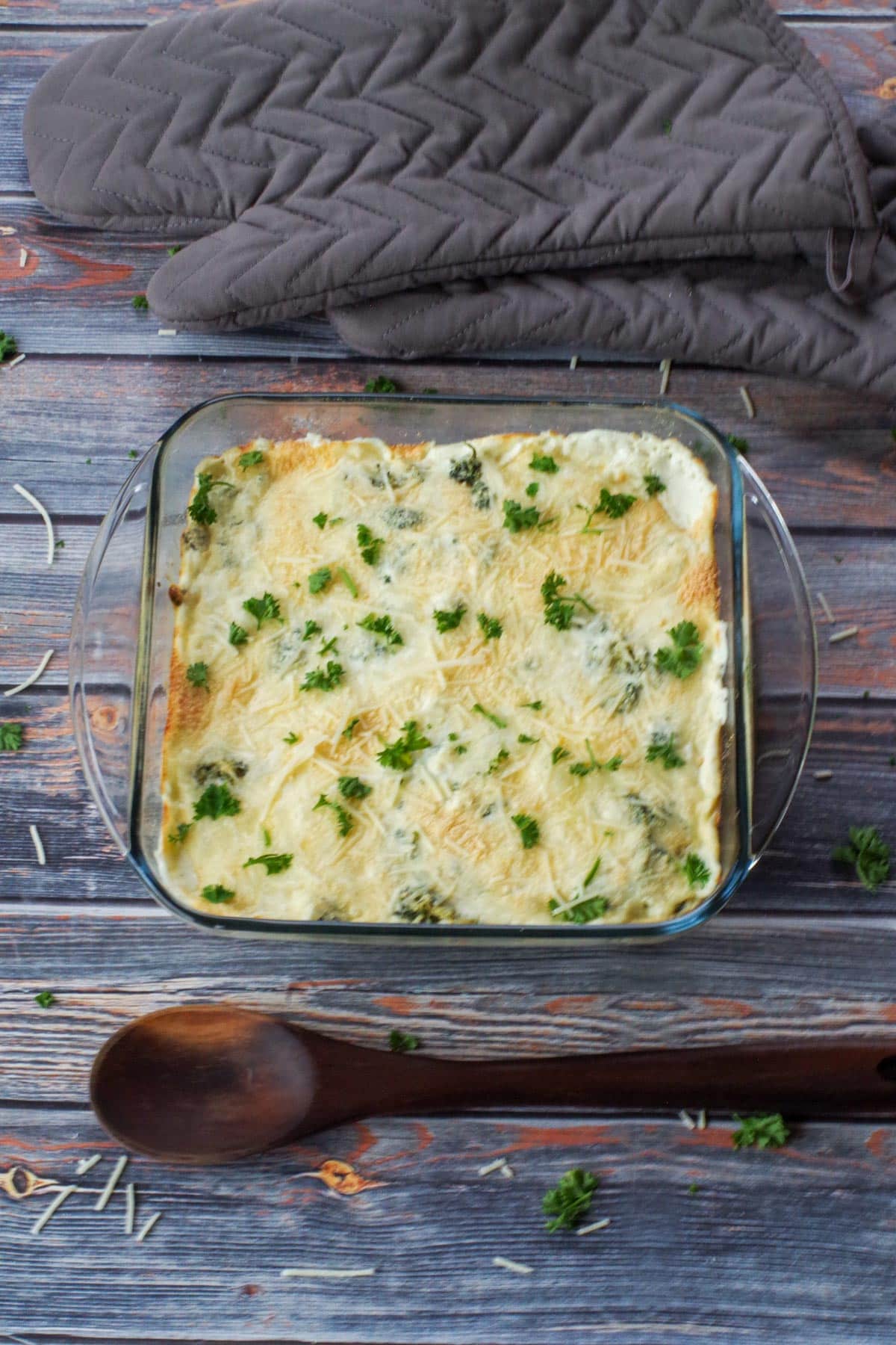 spinach artichoke casserole with wooden spoon below it and grey oven mitts above casserole