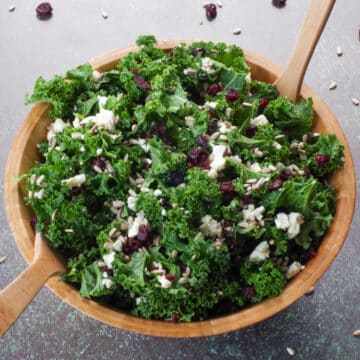 Kale Cranberry and Feta salad in a wooden bowl with wooden utensils and cranberries and sunflower seeds strewn around