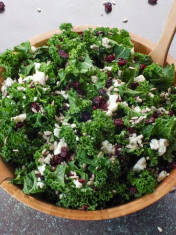Kale Cranberry and Feta salad in a wooden bowl with wooden utensils and cranberries and sunflower seeds strewn around