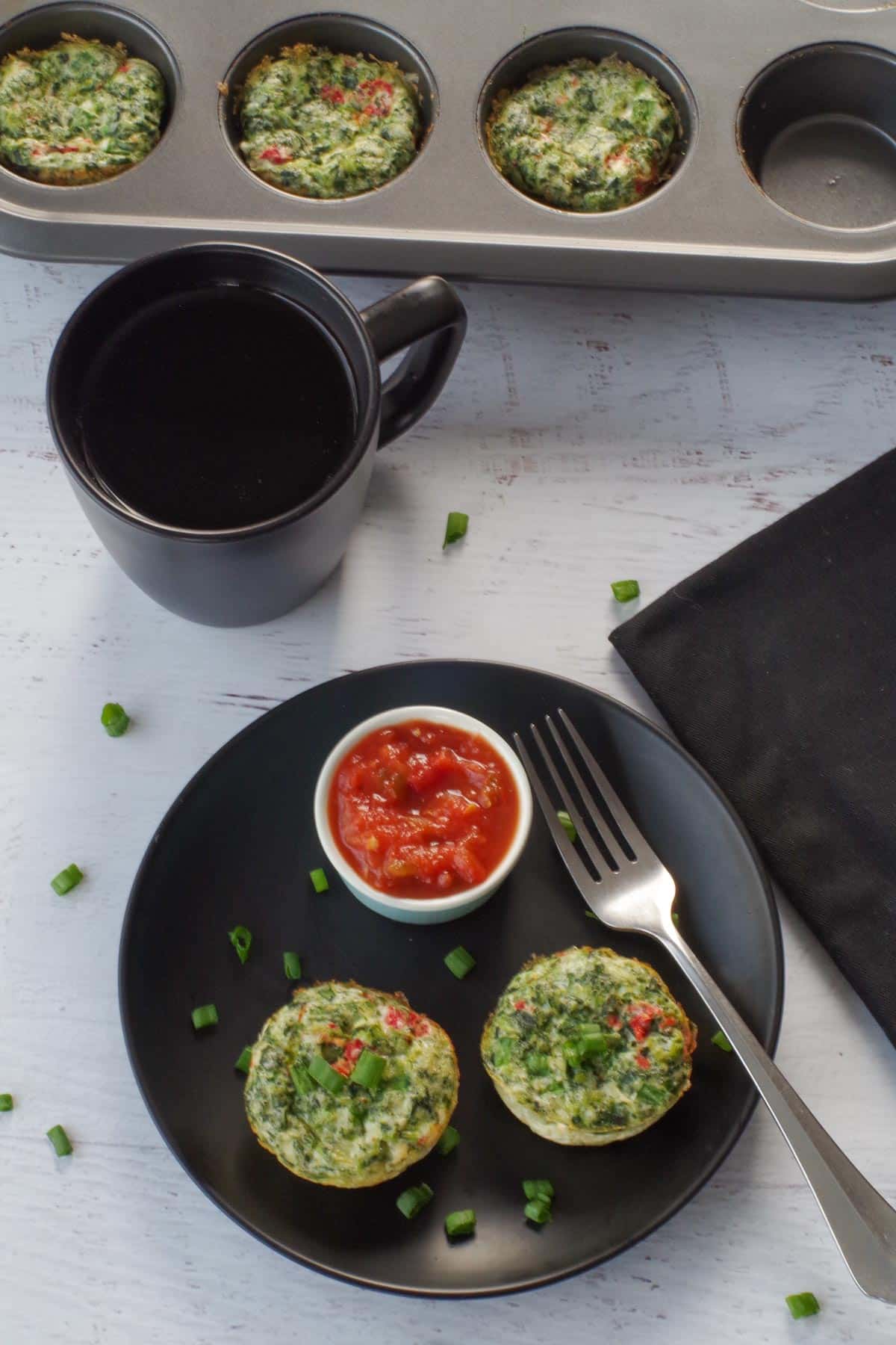 2 egg white bites on a black plate, with salsa in a small dish, a fork on the plate and a muffin tin with more egg bites in the background