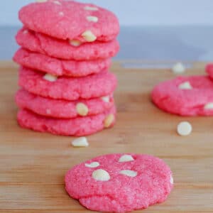 strawberry milkshake cookies on a wood cutting board, with a stack of cookies background and white chocolate chips scattered
