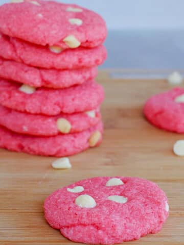 strawberry milkshake cookies on a wood cutting board, with a stack of cookies background and white chocolate chips scattered